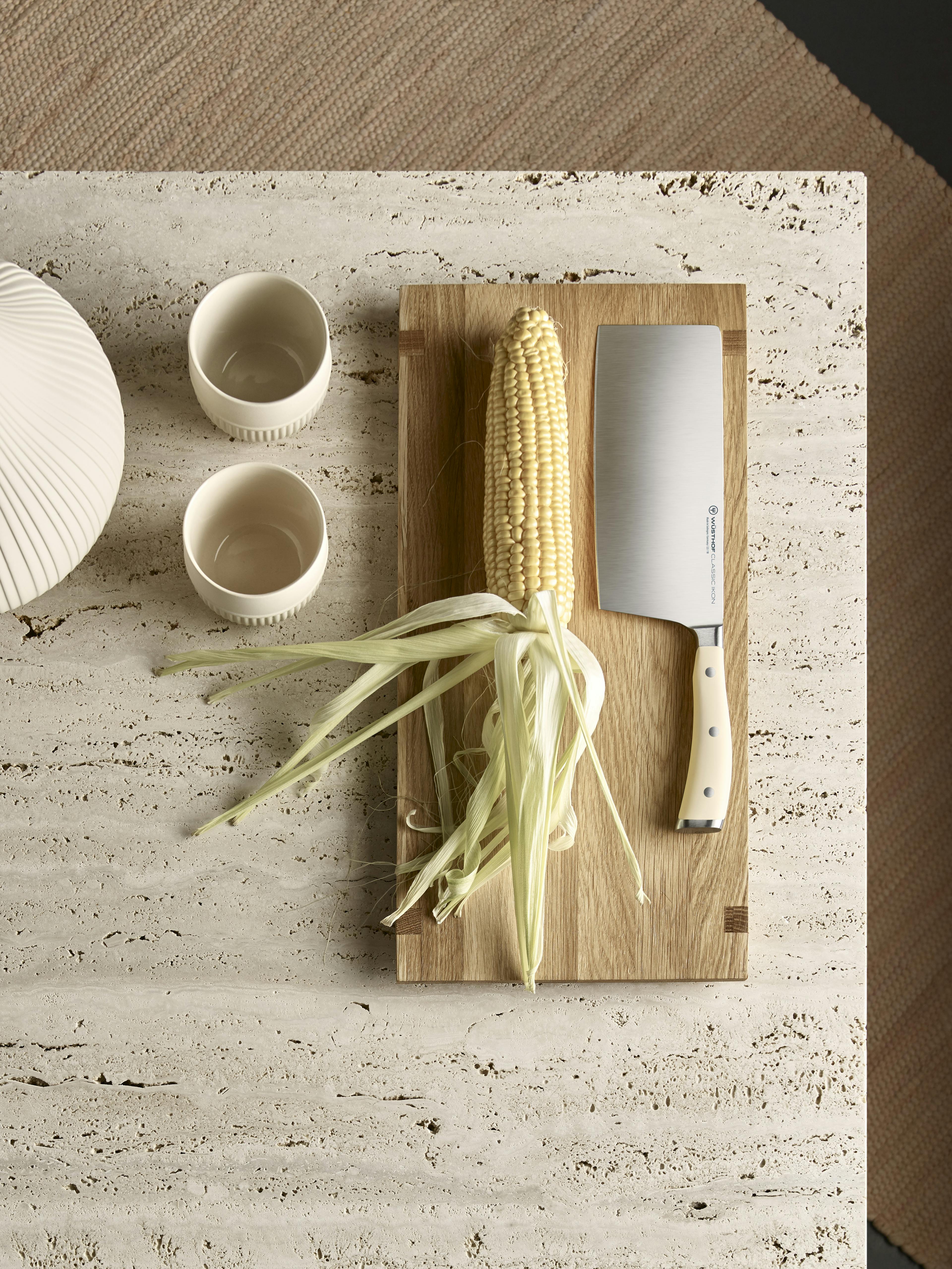 An ear of corn laying on a cutting board next to a WÜSTHOF Classic Ikon Crème Chinese Chef's Knife